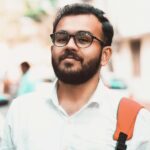 Confident man with glasses and backpack on a sunny urban street with cars in the background.